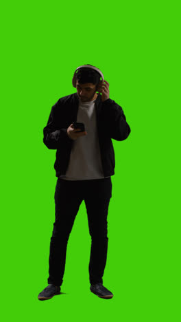 Vertical-Video-Full-Length-Shot-Of-Man-Wearing-Wireless-Headphones-Streaming-Music-From-Mobile-Phone-Against-Green-Screen-2